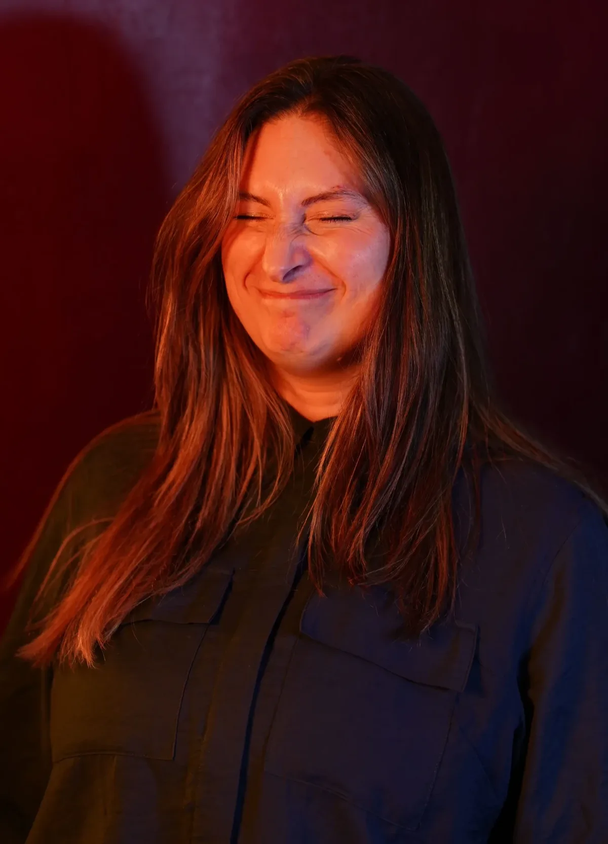 Donna, founder and piercer at The Needle Stack, grimacing playfully under dramatic red and blue studio lighting with a dark red background