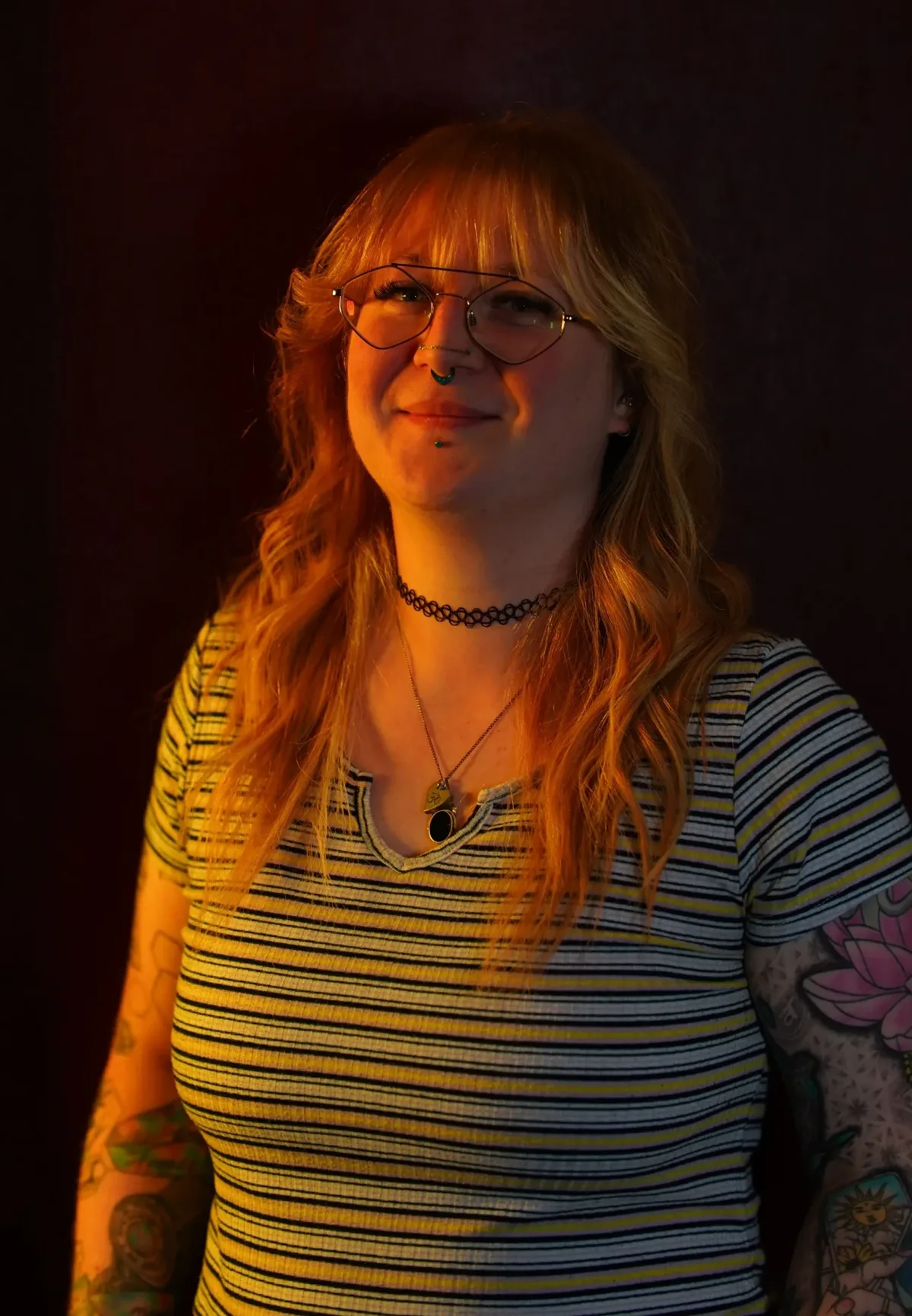 Kim, resident tattoo artist at Requiem Tattoo Shropshire, with strawberry blonde hair, hexagonal glasses and facial piercings, arms covered in colourful tattoos, lit by warm orange studio lighting against a dark background