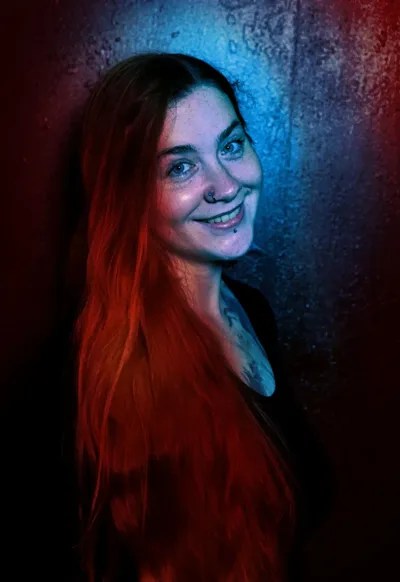 Liz Kuzmin, co-owner and manager of Requiem Tattoo Shropshire, with long red hair and facial piercings, lit by dramatic split red and blue studio lighting